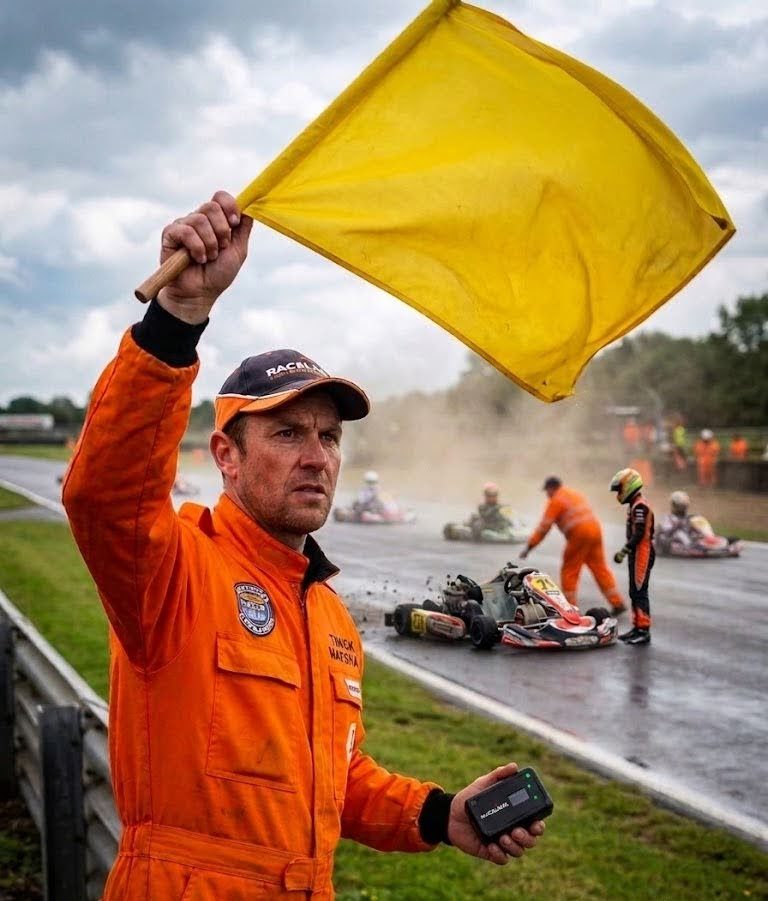 Track marshal raising a yellow flag and holding a RaceLink device at a kart incident